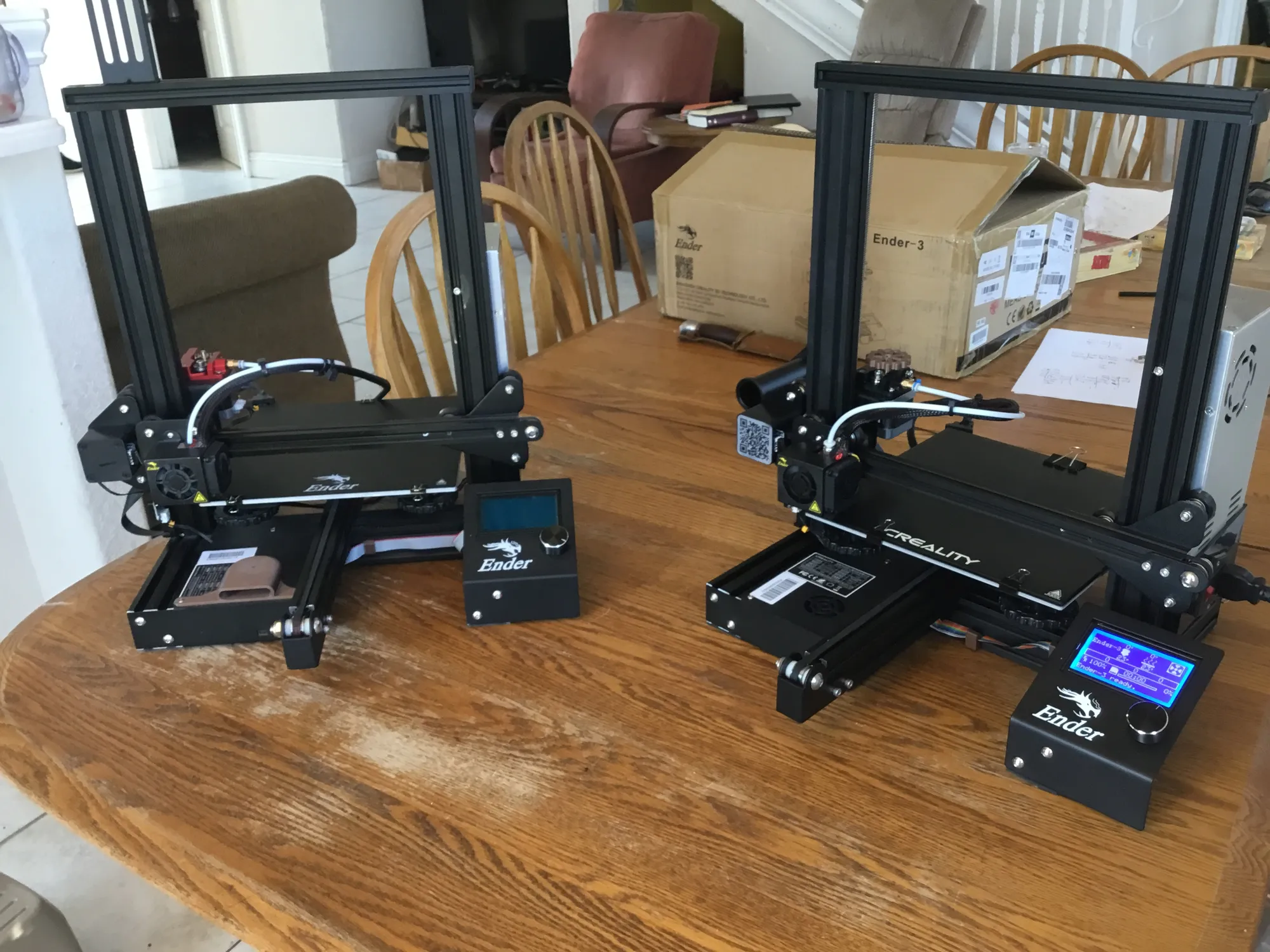 Two black Creality Ender 3D printers on a wooden dining table, the right one with a lit blue screen, an Ender-3 box behind.