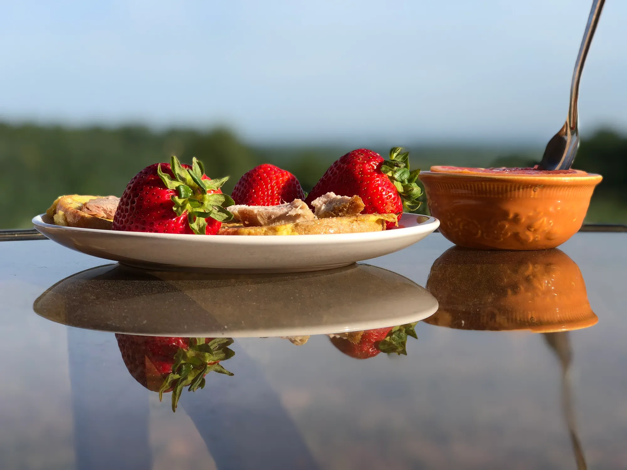 Strawberries, toast and meat on a white plate beside an orange bowl with a spoon, reflected in a glass table outdoors