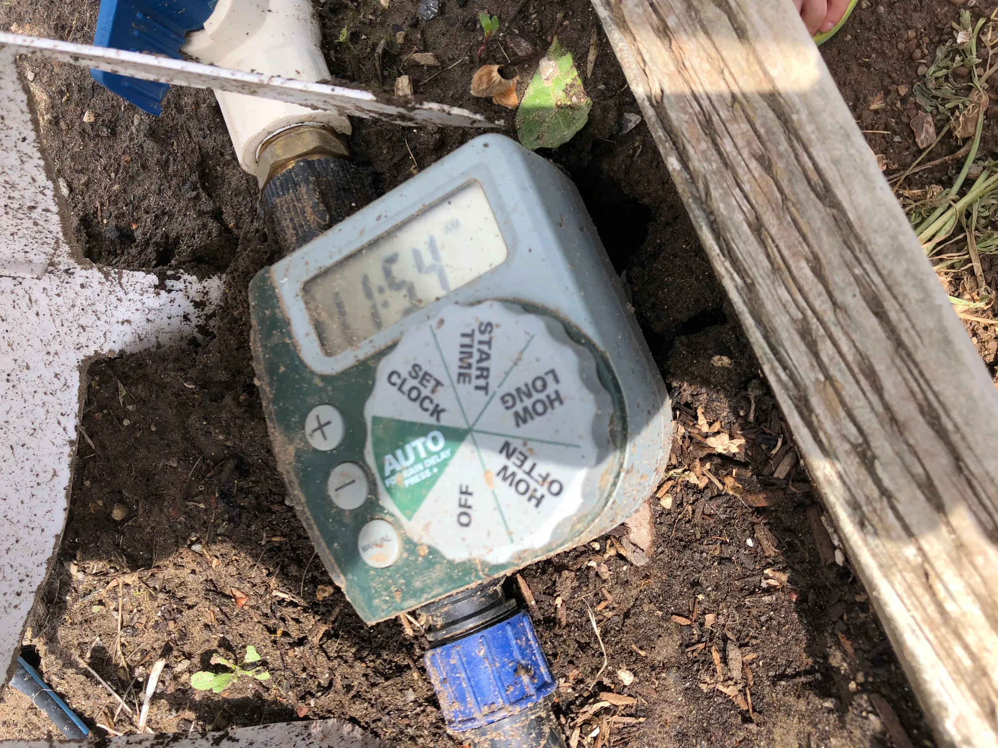 Dirt-covered digital hose timer set to Auto, with a dial and LCD reading 11:54, attached to a pipe in soil beside a wooden