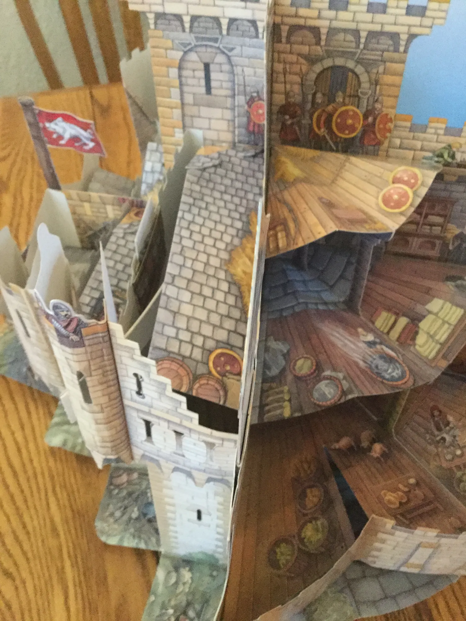 Open pop-up book forming a paper castle with towers, a red flag, guards with shields, and cutaway rooms with storerooms and