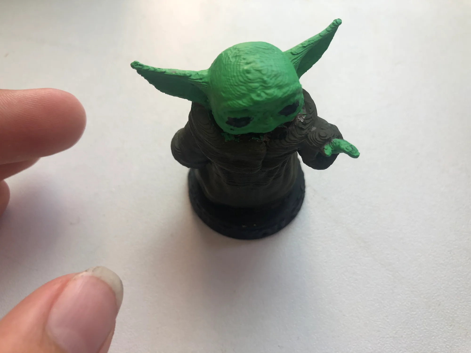 Small 3D-printed Baby Yoda figure painted green with a dark brown robe, reaching out a hand, beside fingertips on a white