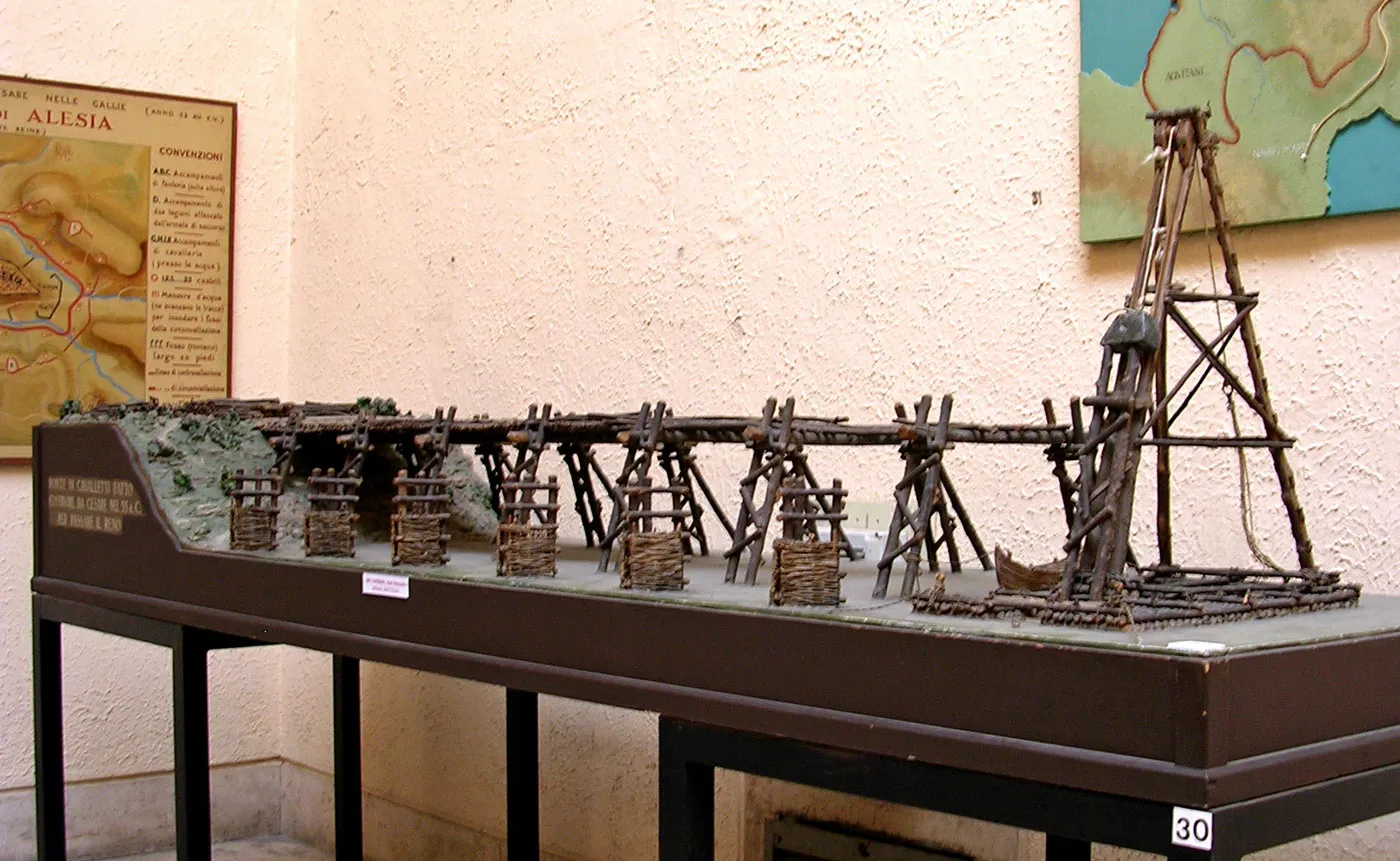 Museum model of a wooden trestle bridge with wicker baskets and a pile-driving tower on a table, maps of Alesia on the wall
